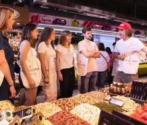 La gente conversa en un mercado, con diversos productos gastronómicos expuestos, como la llamativa "Clase de cocina de paella en Barcelona". Un cartel dice "Sprim Frut" al fondo. - ForTwo - Planes para parejas