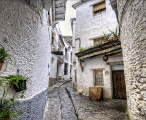 Una estrecha calle adoquinada bordeada de edificios encalados y plantas en macetas, que conduce cuesta arriba y que recuerda a un pueblo de la Alpujarra de Granada que aparece en "Alpujarra: Tour guiado desde Granada". - ForTwo - Planes para parejas