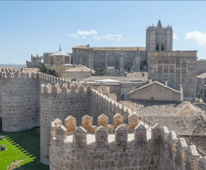 Los muros de piedra medievales de Ávila y una catedral se alzan majestuosos contra el cielo azul claro, presentados en el tour "Ávila y Segovia: Excursión de un día desde Madrid con Entrada al Alcázar de Segovia". - ForTwo - Planes para parejas