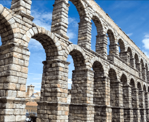 Un histórico acueducto de piedra con múltiples arcos se eleva bajo un cielo azul brillante, enmarcado por edificios de la ciudad, haciendo eco de la grandeza de la arquitectura cerca del Alcázar de Segovia. Experimenta esto en "Ávila y Segovia: Excursión de un día desde Madrid con Entrada al Alcázar de Segovia. - ForTwo - Planes para parejas