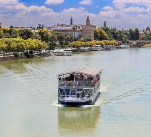 El "Crucero Torre del Oro por el Guadalquivir" ofrece vistas panorámicas de la Torre del Oro y la arquitectura de la ciudad en medio de una vegetación vibrante bajo un cielo azul claro. - ForTwo - Planes para parejas