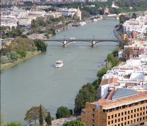 El tranquilo río Guadalquivir se distingue por su puente arqueado, barcos como el Crucero Torre del Oro por el Guadalquivir que pasan suavemente, y edificios urbanos a ambas orillas. La icónica Torre del Oro realza el paisaje con su encanto histórico. - ForTwo - Planes para parejas