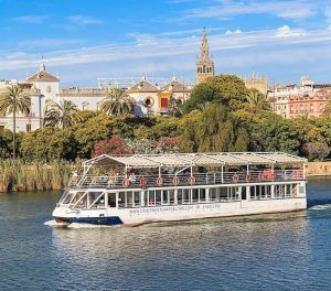 El Crucero Torre del Oro por el Guadalquivir se desliza elegantemente sobre el río Guadalquivir, rodeado de palmeras ondulantes y edificios históricos, con la icónica Torre del Oro al fondo bajo un cielo azul claro. - ForTwo - Planes para parejas
