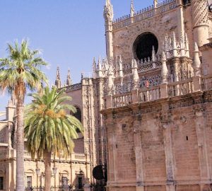 La majestuosa Giralda se alza sobre una catedral gótica con intrincadas obras de piedra y palmeras bajo un cielo azul despejado. Disfrute de una visita inolvidable a la icónica Catedral de Sevilla con la "Entrada para la Catedral de Sevilla y la Giralda (sin colas)" para un acceso rápido y sin complicaciones. - ForTwo - Planes para parejas