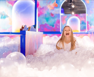 Una persona se sienta alegremente en una piscina de bolas llena de pelotas blancas, rodeada de coloridas paredes de ensueño en el Museo Sweet Space de Madrid. ¡Reserva tus entradas al Museo Sweet Space de Madrid para esta fantástica aventura!. - ForTwo - Planes para parejas