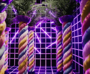 Columnas retorcidas y palmeras bañadas por luces de neón en una sala similar al Museo Sweet Space de Madrid, presentan una pared de cuadrícula en blanco y negro realzada por luces púrpuras para crear un ambiente de otro mundo. - ForTwo - Planes para parejas