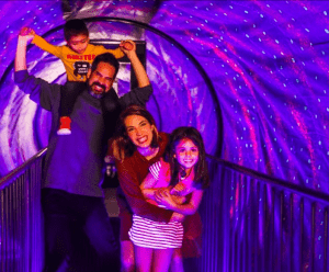 Una familia de cuatro sonríe y juega en un vibrante y colorido túnel iluminado en el Museo de las Ilusiones de Madrid, creando recuerdos inolvidables con Entradas al Museo de las Ilusiones de Madrid. - ForTwo - Planes para parejas