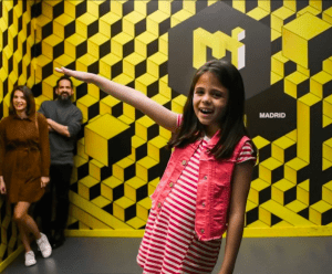 Una niña posa juguetonamente con un vestido de rayas en la sala geométrica amarilla del Museo de las Ilusiones de Madrid, mientras dos adultos sonríen al fondo. - ForTwo - Planes para parejas