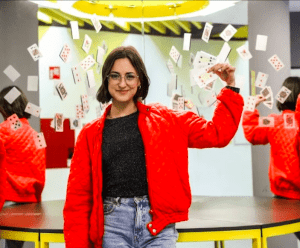 Una persona con chaqueta roja sonríe, sosteniendo naipes mientras otros flotan a su alrededor. El espejo refleja esta escena lúdica, evocando la experiencia de "Entradas al Museo de las Ilusiones de Madrid". - ForTwo - Planes para parejas