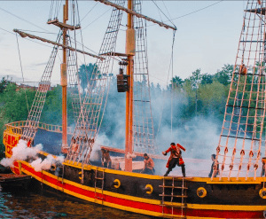 Un barco pirata con una tripulación animada y velas coloridas surca la niebla, con sus cañones humeantes. Con el fondo boscoso, parece como si hubieran emergido de Agua Mágica, trayendo historias de maravillas similares a las del Acuario de Sevilla descritas en Entradas para Agua Mágica y Acuario de Sevilla. - ForTwo - Planes para parejas