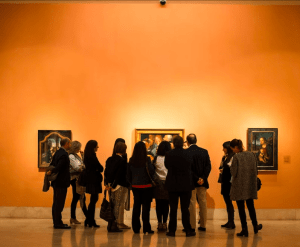 Un grupo animado aprecia las obras de arte en una vibrante pared de galería de color naranja en el Museo Thyssen, mezclando cultura y creatividad durante el evento "Entradas para Brunch y Arte en el Museo Thyssen". - ForTwo - Planes para parejas