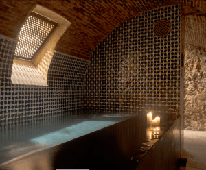 Experimente la lujosa "Experiencia Hammam Tradicional - Baño Turco en Madrid", donde una vela encendida y paredes de azulejos crean un ambiente de tranquilidad. Una ventana arqueada ilumina este espacio sereno, evocando la elegancia atemporal de los mejores retiros de Madrid. - ForTwo - Planes para parejas