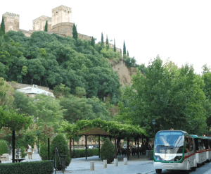 Los autobuses están estacionados cerca de frondosos árboles con el histórico castillo de Granada en una colina al fondo, mientras que las Entradas para Tren turístico en Granada ofrecen una exploración más cercana de este encantador paisaje. - ForTwo - Planes para parejas