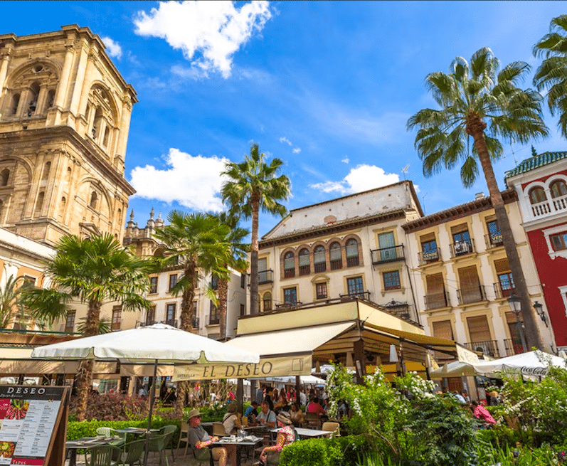 Un café al aire libre en una plaza vibrante con altas palmeras y edificios históricos, en medio del alegre zumbido del tren turístico en Granada, que pasa bajo un cielo azul infinito. - ForTwo - Planes para parejas