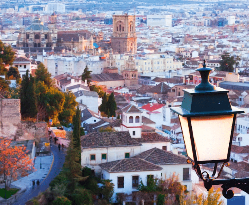 Una vista panorámica de Granada al atardecer, con edificios históricos bajo una luz dorada, una encantadora farola con suaves sombras y montañas a lo lejos. Esta vista es tan atemporal como un billete de "Entradas para Tren Turístico en Granada" a su pasado histórico. - ForTwo - Planes para parejas
