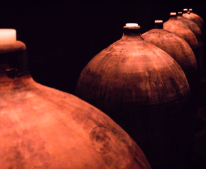Una hilera de grandes tinajas de barro rústicas con tapa, tenuemente iluminadas sobre un fondo oscuro, evoca el ambiente de una "Excursión y Cata de Vinos de la Ribera del Duero desde Madrid. - ForTwo - Planes para parejas