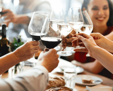 Personas levantando copas de vino en un brindis en una mesa de comedor con botellas y pan de Excursión y Cata de Vinos de la Ribera del Duero desde Madrid visibles, que recuerdan a una elegante cata de vinos. - ForTwo - Planes para parejas