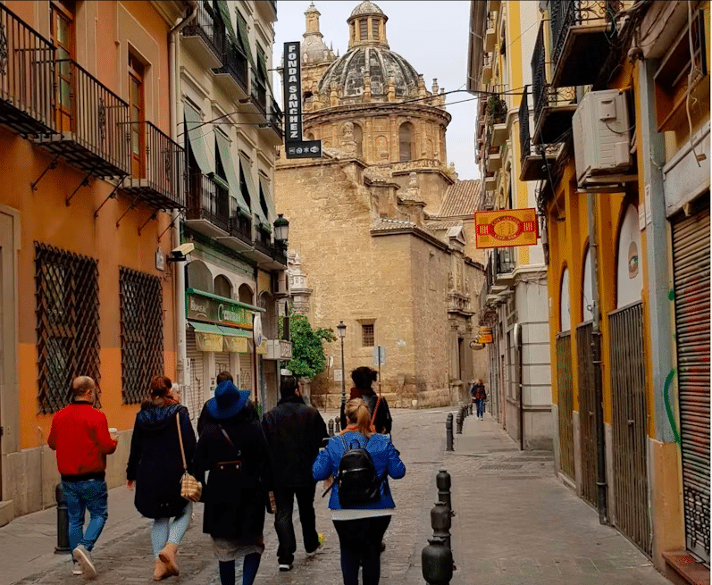 Caminar por una estrecha calle europea entre edificios históricos con la cúpula de una iglesia al fondo se siente como estar en el "Federico García Lorca: Tour guiado por Granada", donde los ecos de la historia llenan el aire. - ForTwo - Planes para parejas