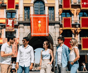 Un grupo de personas sonríe y charla frente a un arco decorado con pancartas rojas y un emblema de cáliz, haciéndose eco de la vibrante escena cultural de "Madrid y Toledo: Excursión guiada de un día y Museo del Prado". - ForTwo - Planes para parejas