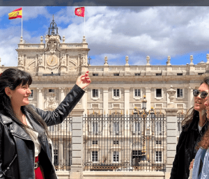 Dos mujeres sonriendo frente a un edificio histórico de Madrid con banderas españolas, bajo un cielo parcialmente nublado, disfrutando de sus Entradas para Madrid: Visita de día completo con entrada rápida al Museo del Prado y al Palacio Real. - ForTwo - Planes para parejas