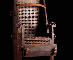 Una silla de tortura de madera, con púas y ataduras, se yergue siniestramente sobre un fondo negro: una reliquia inquietante que recuerda las escalofriantes exhibiciones de Entradas sin colas al Palacio de los Olvidados. - ForTwo - Planes para parejas