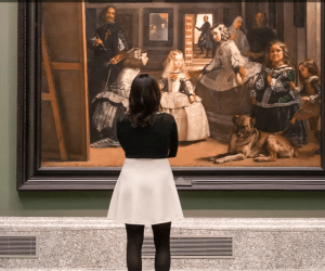 En el Museo del Prado, una mujer vestida de blanco y negro examina atentamente una gran pintura enmarcada como parte de su experiencia de recorrido de un día completo con entrada rápida al icónico museo y al Palacio Real. - ForTwo - Planes para parejas