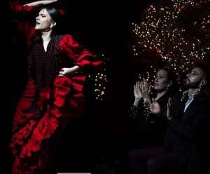 Una bailarina con un vestido rojo interpreta flamenco en Tablao Flamenco, hipnotizando al público con entradas vibrantes mientras dos personas con trajes oscuros observan y aplauden bajo las luces centelleantes de Torres Bermejas. - ForTwo - Planes para parejas