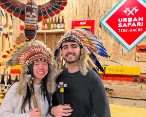Dos personas con tocados de plumas sostienen hachas en una habitación de temática rústica, disfrutando de una emocionante "Tiro con Hacha sesión 1 hora" bajo el cartel de Urban Safari. - ForTwo - Planes para parejas