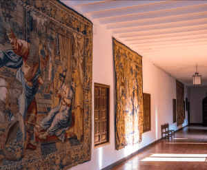 Un pasillo iluminado por el sol con tapices ornamentados en paredes blancas, bancos de madera y un suelo de baldosas evoca el encanto histórico de la "Visita la Abadía del Sacromonte + Museo Cuevas del Sacromonte". - ForTwo - Planes para parejas