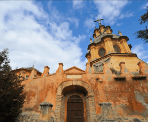 Una iglesia de estilo mediterráneo de color naranja con una cruz se eleva majestuosamente contra un cielo azul lleno de nubes, capturando la serena belleza que se encuentra cerca de la Abadía del Sacromonte y el Museo Cuevas del Sacromonte. - ForTwo - Planes para parejas