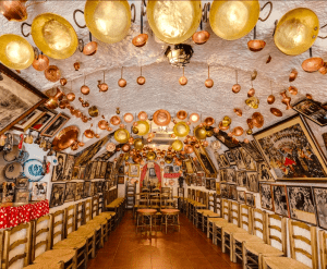 Visita una sala inspirada en el Sacromonte a través de la "Visita la Abadía del Sacromonte + Museo Cuevas del Sacromonte", que cuenta con sillas rústicas, mesas y paredes adornadas con fotografías enmarcadas y macetas de latón colgantes. - ForTwo - Planes para parejas