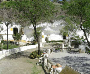 Una casa cueva con un camino ajardinado, rodeada de árboles y flores, se encuentra cerca del cautivador Museo Cuevas del Sacromonte de la experiencia "Visita la Abadía del Sacromonte + Museo Cuevas del Sacromonte". - ForTwo - Planes para parejas