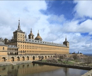 Un gran edificio histórico con torres y un estanque, que recuerda la grandeza del Monasterio del Escorial, se alza contra un cielo azul, invitando a todos a imaginar las Entradas al Monasterio del Escorial: Visita guiada que dan vida a la historia. - ForTwo - Planes para parejas