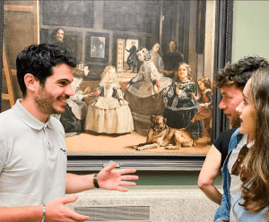 Tres personas conversan y sonríen frente a un cuadro clásico, capturando la esencia del rico patrimonio artístico de Madrid, que recuerda a la exploración de tesoros con "Entradas para Madrid: Visita de día completo con entrada rápida al Museo del Prado y al Palacio Real. - ForTwo - Planes para parejas