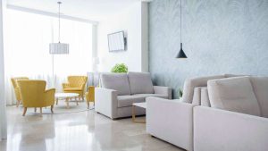 Una suite de GCE Hoteles ofrece una vida moderna con sofás beige, sillones amarillos, paredes gris claro, TV, iluminación colgante y luz natural, ideal para relajarse después de una escapada romántica en Cártama. - ForTwo - Planes para parejas