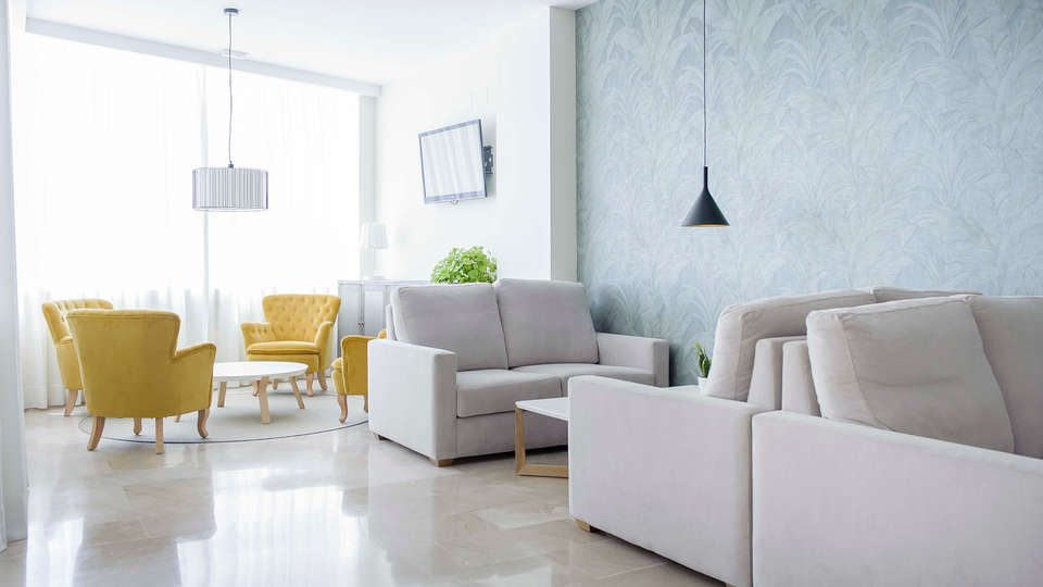 Una suite de GCE Hoteles ofrece una vida moderna con sofás beige, sillones amarillos, paredes gris claro, TV, iluminación colgante y luz natural, ideal para relajarse después de una escapada romántica en Cártama. - ForTwo - Planes para parejas