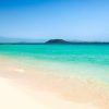 Las cristalinas olas del mar turquesa acarician suavemente la playa de arena de Fuerteventura, con una isla a lo lejos recortada bajo un cielo azul, ofreciendo romance y relajación. Disfruta de una "Escapada en pareja en un resort todo incluido en Fuerteventura". - ForTwo - Planes para parejas