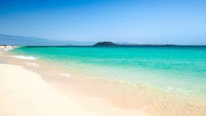 Las cristalinas olas del mar turquesa acarician suavemente la playa de arena de Fuerteventura, con una isla a lo lejos recortada bajo un cielo azul, ofreciendo romance y relajación. Disfruta de una "Escapada en pareja en un resort todo incluido en Fuerteventura". - ForTwo - Planes para parejas