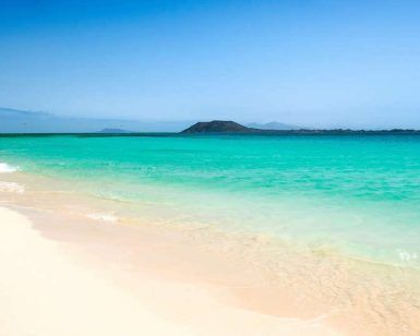 Las cristalinas olas del mar turquesa acarician suavemente la playa de arena de Fuerteventura, con una isla a lo lejos recortada bajo un cielo azul, ofreciendo romance y relajación. Disfruta de una "Escapada en pareja en un resort todo incluido en Fuerteventura". - ForTwo - Planes para parejas
