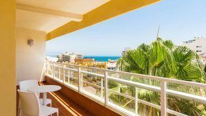Disfrute de un soleado balcón con dos sillas blancas y una mesa en el Hotel Roc Flamingo. Con vistas a los edificios y al océano, es el lugar perfecto para una escapada a Torremolinos. Una gran palmera contribuye a la tranquilidad del lugar. - ForTwo - Planes para parejas