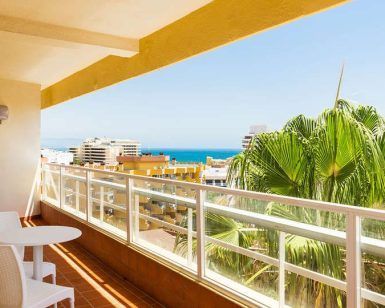 Disfrute de un soleado balcón con dos sillas blancas y una mesa en el Hotel Roc Flamingo. Con vistas a los edificios y al océano, es el lugar perfecto para una escapada a Torremolinos. Una gran palmera contribuye a la tranquilidad del lugar. - ForTwo - Planes para parejas