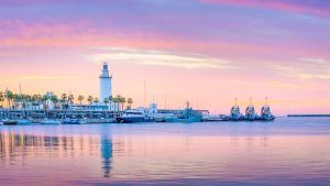 La serena puesta de sol en el puerto, con un faro y palmeras, enmarcada por barcos contra un cielo rosa y morado, es ideal para tu "Escapada a un apartahotel de Málaga: Soho Boutique Museo". Imagina disfrutar de esta vista después de explorar la vibrante ciudad. - ForTwo - Planes para parejas