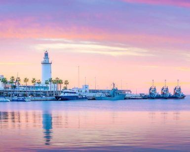 La serena puesta de sol en el puerto, con un faro y palmeras, enmarcada por barcos contra un cielo rosa y morado, es ideal para tu "Escapada a un apartahotel de Málaga: Soho Boutique Museo". Imagina disfrutar de esta vista después de explorar la vibrante ciudad. - ForTwo - Planes para parejas