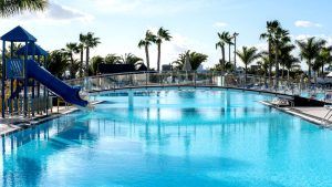Descubre el encanto de Escapada, un resort en Lanzarote, en Playa Blanca: Hotel THB Tropical Island. Relájate en una piscina al aire libre con tobogán azul, palmeras y un puente panorámico, todo bajo un cielo despejado. - ForTwo - Planes para parejas