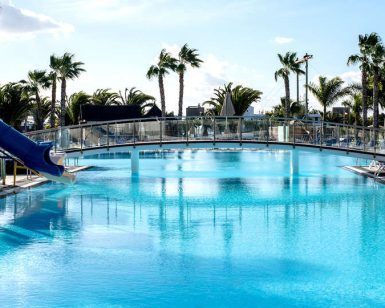Descubre el encanto de Escapada, un resort en Lanzarote, en Playa Blanca: Hotel THB Tropical Island. Relájate en una piscina al aire libre con tobogán azul, palmeras y un puente panorámico, todo bajo un cielo despejado. - ForTwo - Planes para parejas