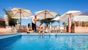 Un hombre y una mujer se relajan junto a la piscina del Hotel Globales Acuario, disfrutando de unas bebidas con un camarero cerca. Las sombrillas ofrecen sombra bajo el sol radiante: una escapada ideal a Tenerife. - ForTwo - Planes para parejas