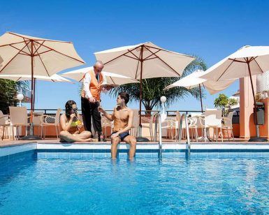 Un hombre y una mujer se relajan junto a la piscina del Hotel Globales Acuario, disfrutando de unas bebidas con un camarero cerca. Las sombrillas ofrecen sombra bajo el sol radiante: una escapada ideal a Tenerife. - ForTwo - Planes para parejas