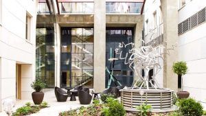 Un moderno patio en Budapest exhibe una escultura abstracta de metal, plantas en macetas y sillas redondas con un fondo de escalera de vidrio, ideal para una escapada romántica en el Hotel Continental 4 estrellas (Budapest). - ForTwo - Planes para parejas