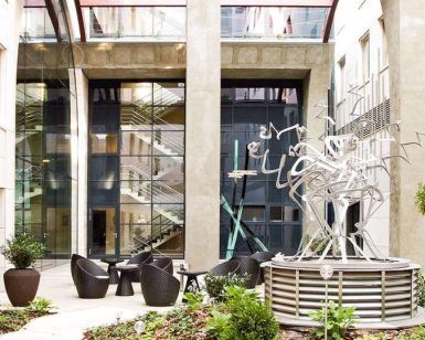 Un moderno patio en Budapest exhibe una escultura abstracta de metal, plantas en macetas y sillas redondas con un fondo de escalera de vidrio, ideal para una escapada romántica en el Hotel Continental 4 estrellas (Budapest). - ForTwo - Planes para parejas
