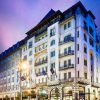 Una vista de la calle del gran y ornamentado hotel Novotel Budapest Centrum al anochecer, con luces brillantes que proyectan un resplandor romántico bajo el cielo púrpura de Budapest. - ForTwo - Planes para parejas
